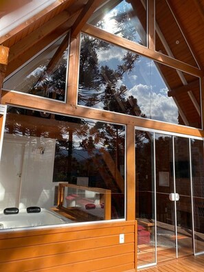A modern wooden cabin with large glass windows, a hot tub, and a view of trees.