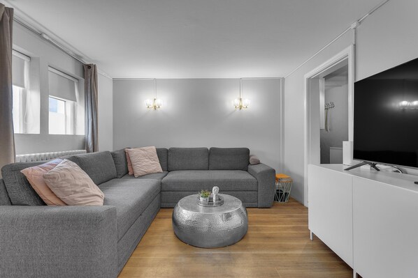 Un salon moderne avec un canapé d’angle gris, une table basse ronde et un téléviseur à écran plat posé sur un meuble blanc.