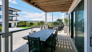 A covered outdoor area with a wooden table and chairs, a hanging chair, and a view of a residential building across the street. - Experience Ocean and Pier Views, Close proximity to the Sand, at this Luxury Bea (Wrightsville Beach)