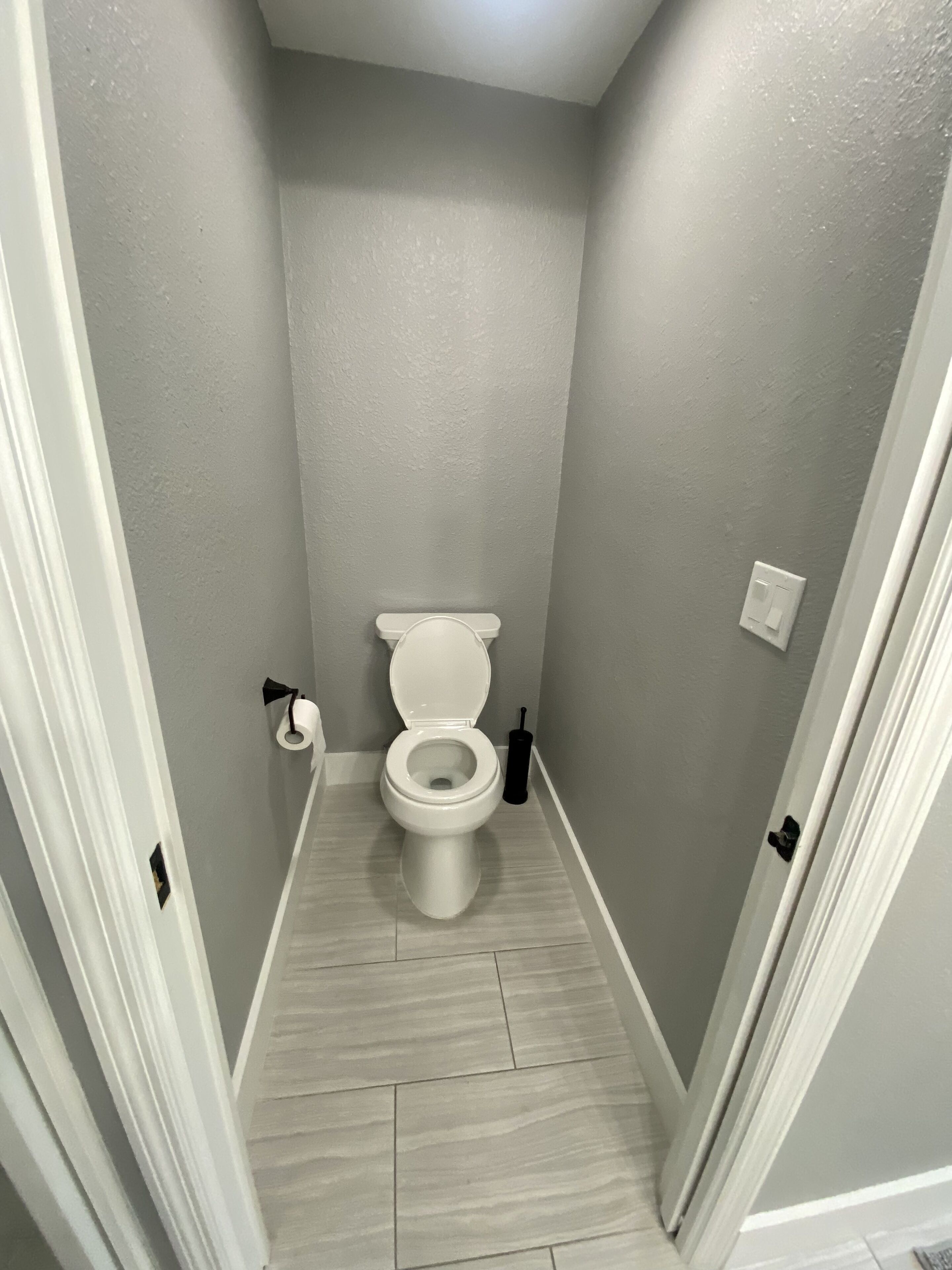 Un baño estrecho con inodoro, un dispensador de papel higiénico y un cepillo de limpieza negro.