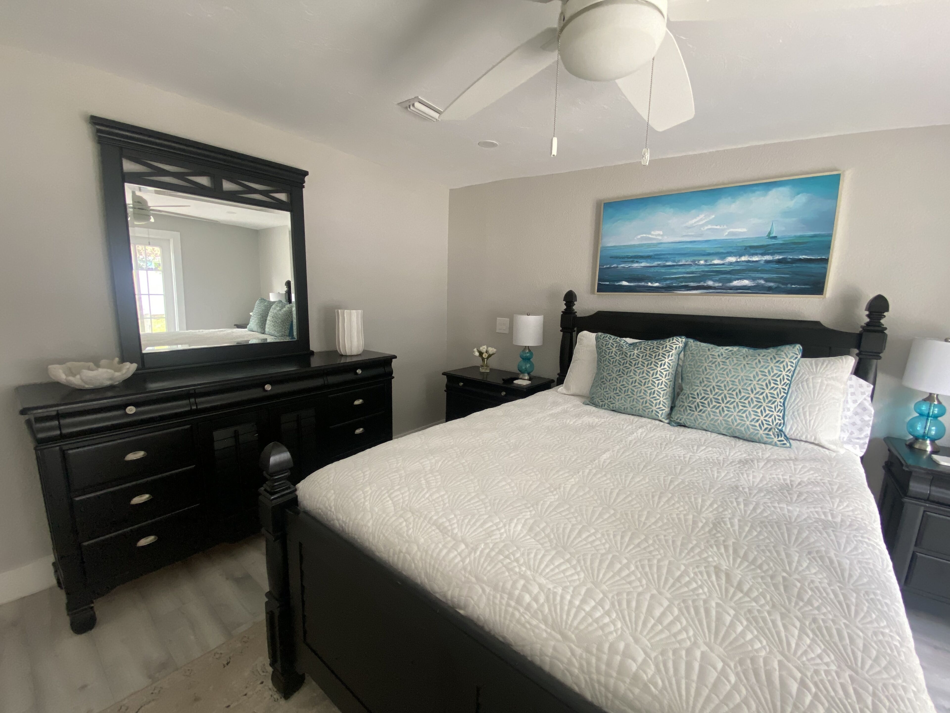 A bedroom with a large bed, a dresser, a nightstand, a ceiling fan, and a painting of the ocean.