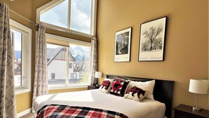 Ein Schlafzimmer mit einem großen Bett, zwei gerahmten Bildern an der Wand, einem Fenster mit Vorhängen und Blick auf einen Berg.