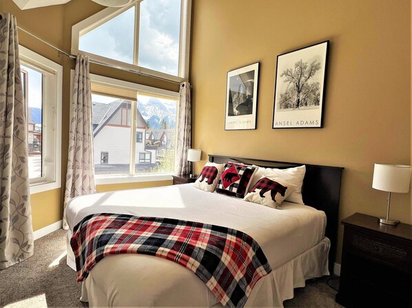 A bedroom with a large bed, two framed pictures on the wall, a window with curtains, and a view of a mountain.