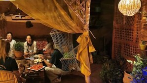 Un groupe de personnes déjeune en plein air, dans un cadre rustique agrémenté d’un hamac et de plantes en pot.