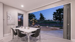 Un moderno comedor con una mesa preparada para cuatro, un amplio ventanal con vista a una cancha de tenis y un puerta corrediza de vidrio que da a un patio exterior.
