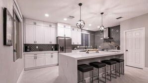 Una cocina moderna con armarios blancos, una isla grande con taburetes de bar, electrodomésticos de acero inoxidable e iluminación colgante.
