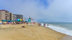 Una playa con gente tomando el sol y nadando, sombrillas y edificios altos al fondo.
