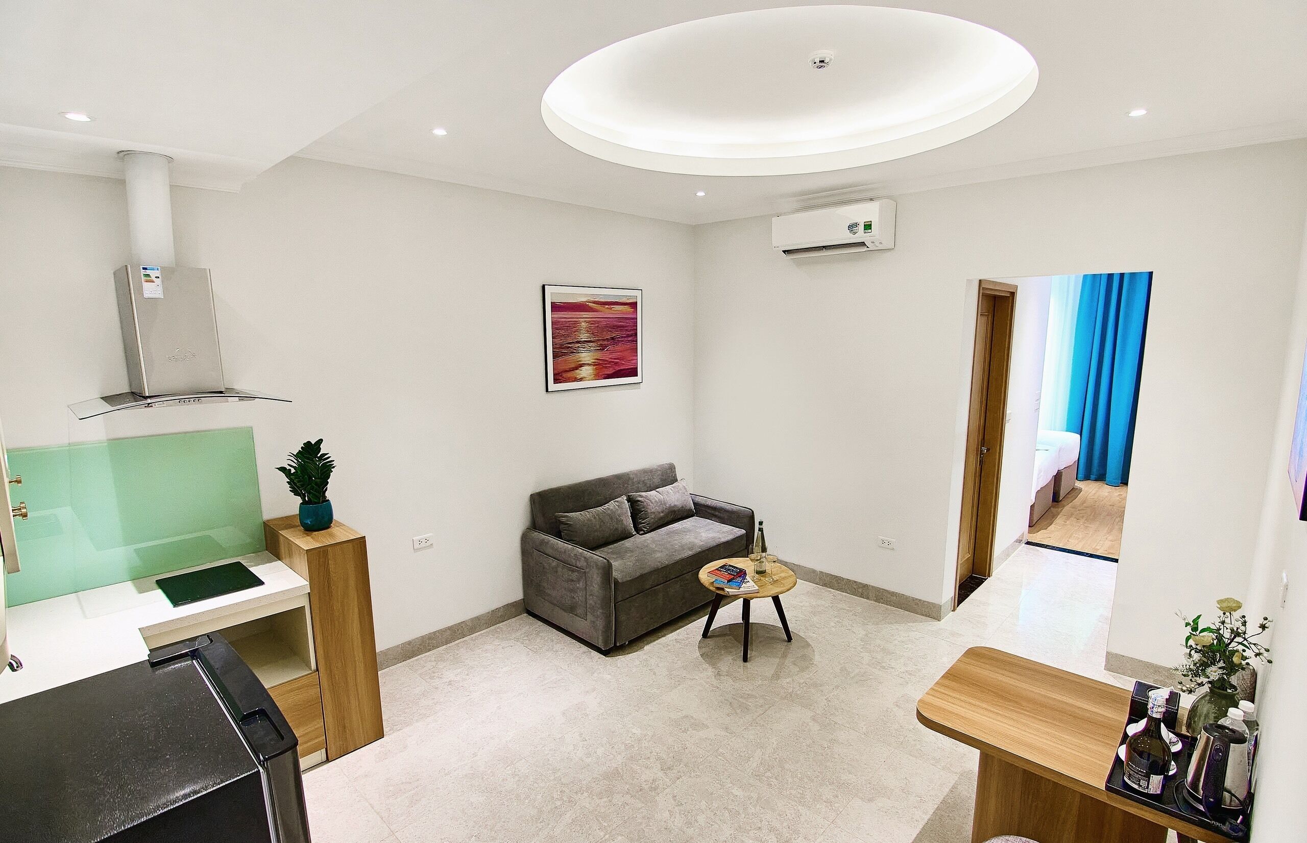 A modern living room with a grey sofa, a round coffee table, a small side table, and a kitchen area with a green countertop and a white stove.