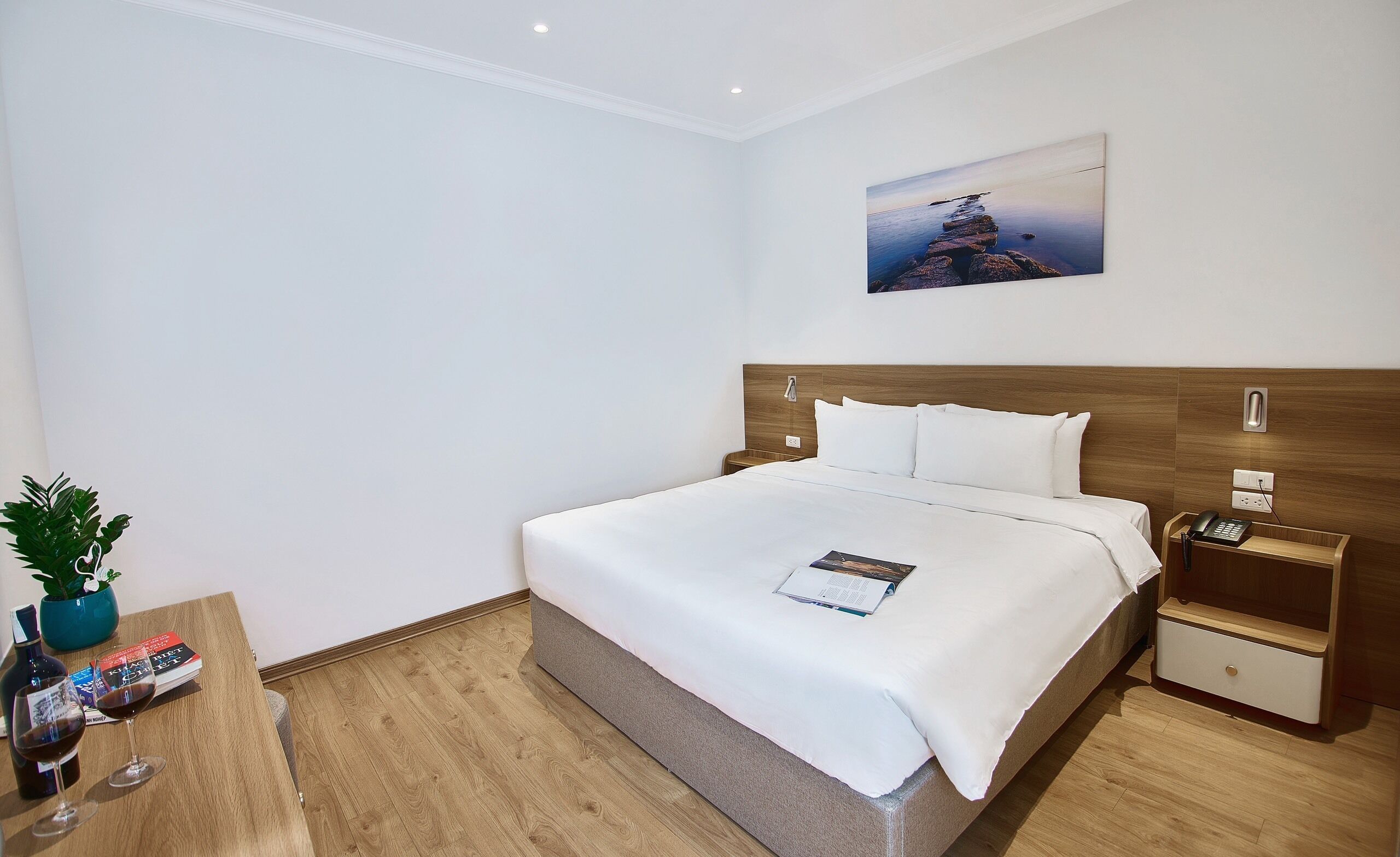 A hotel room with a large bed, a bedside table with a phone, a small nightstand, a wall-mounted light, a painting, and a potted plant.
