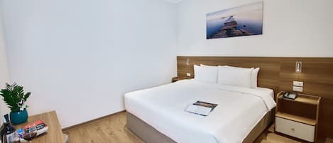A hotel room with a large bed, a bedside table with a phone, a small nightstand, a wall-mounted light, a painting, and a potted plant.