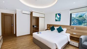 A modern hotel room with a large bed, a wooden wardrobe, a built-in shelf, and a window with a view of greenery.