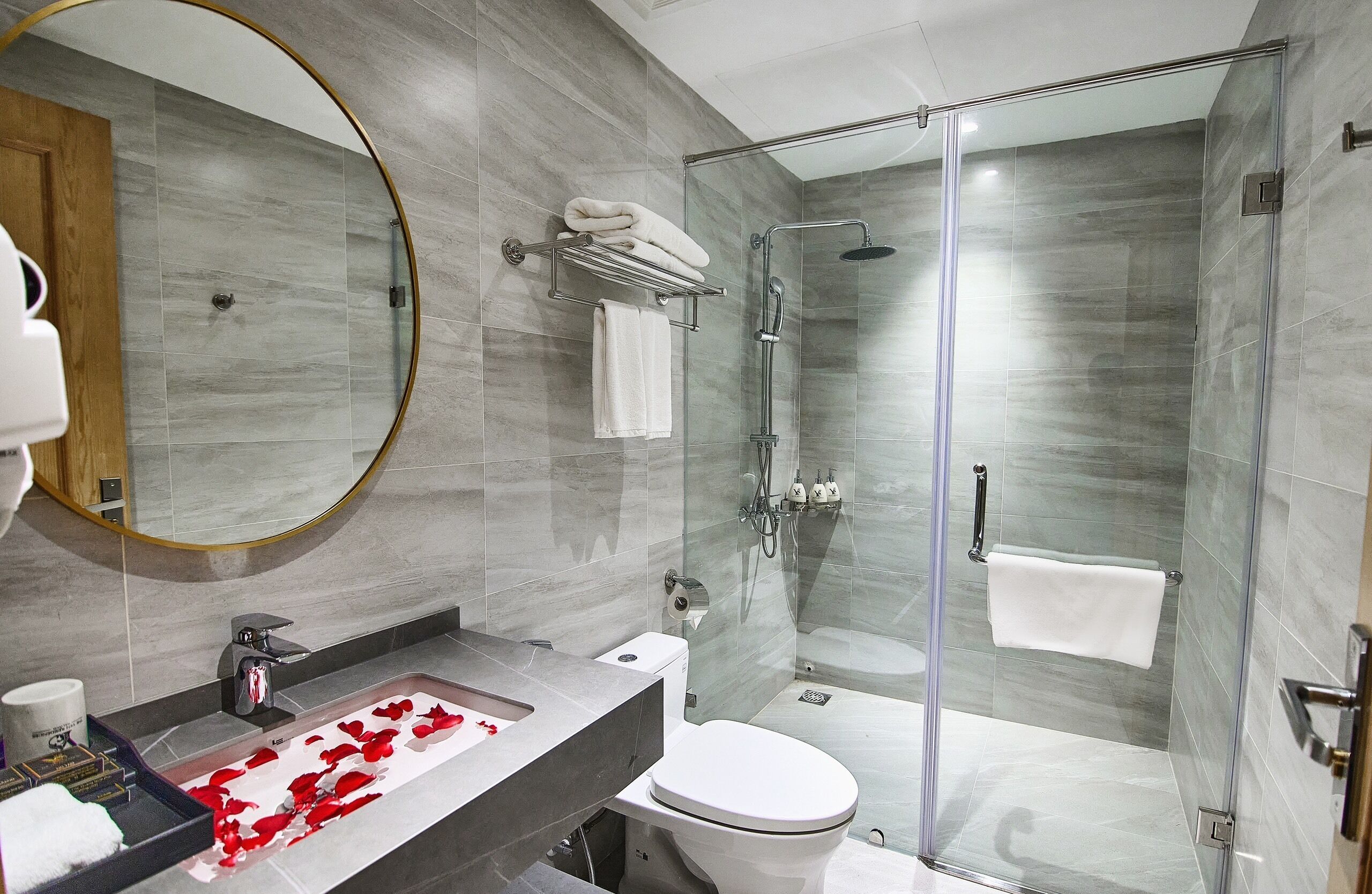A modern bathroom with a glass-enclosed shower, a marble-tiled wall, a round mirror, and a sink with a towel rack.