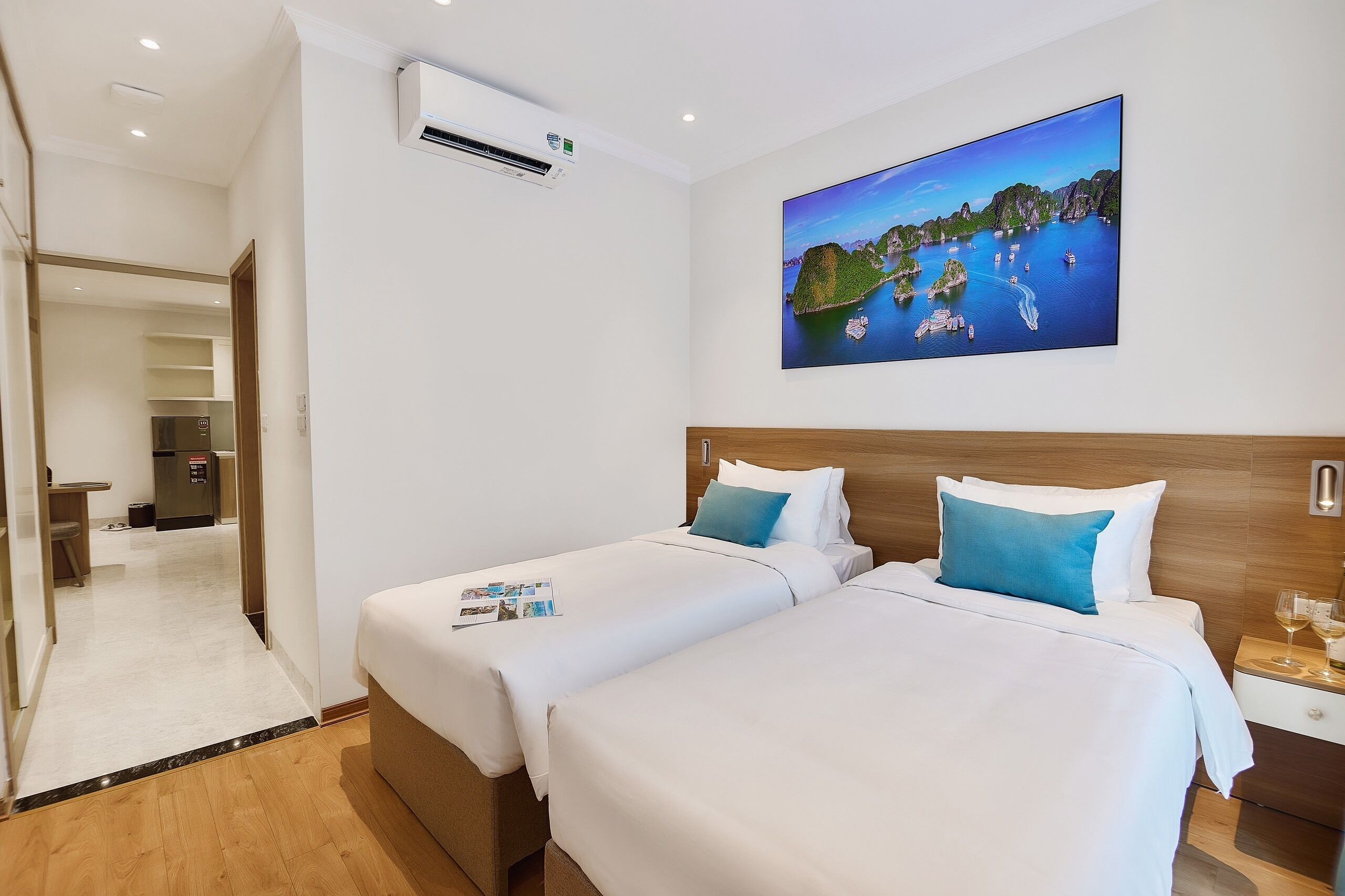 A hotel room with two beds, a painting of a coastal scene, a wooden headboard, and a nightstand with glasses.