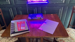 Een houten tafel met een neonbord waarop 'Welcome' staat, een notitieboekje en papieren.