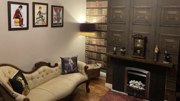 Een woonkamer met een beige bank, een open haard, een boekenkast en lijstwerken aan de muur.