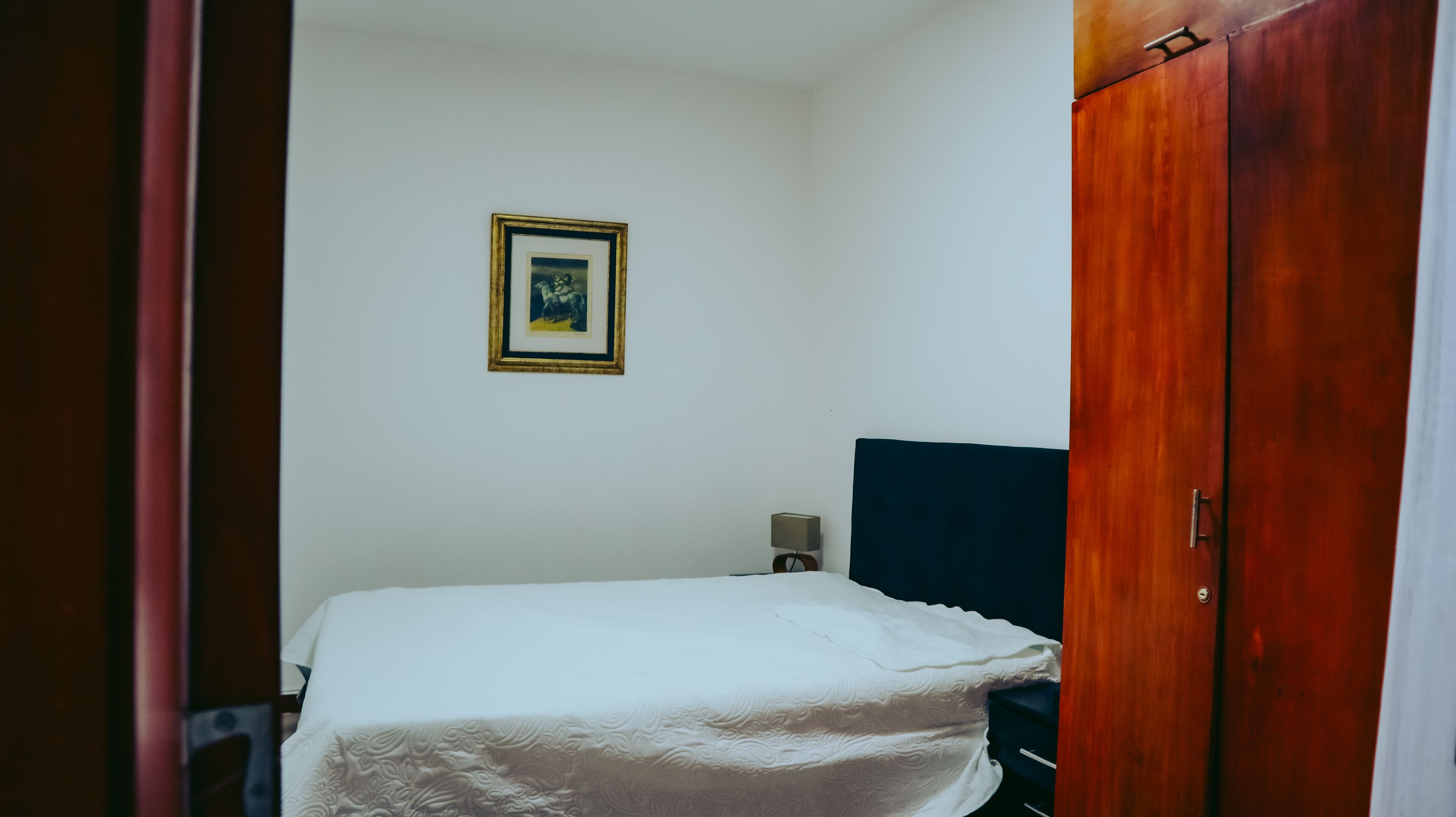 Une petite chambre avec un lit simple, une armoire en bois, un tableau encadré au mur et une table de chevet avec une lampe.