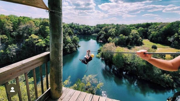 Eine Zipline, auf der eine Person in der Luft über einem von Grünflächen umgebenen Fluss schwebt.