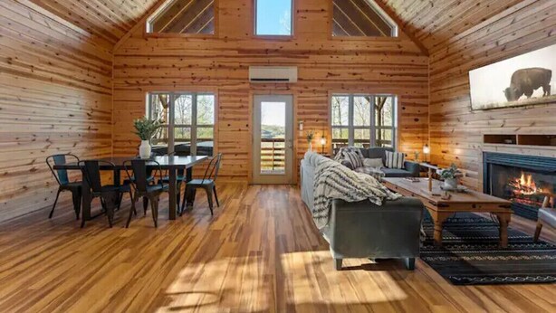 L’intérieur d’une cabane en rondins comprend un coin repas, un canapé, une cheminée et de grandes fenêtres.
