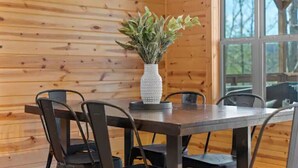 L’intérieur d’un chalet en bois comprend une table à manger et des chaises, un vase blanc à texture particulière agrémenté de feuillage vert, et une grande fenêtre donnant sur un paysage arboré.