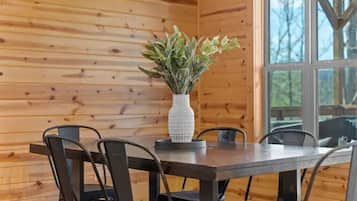 A wooden cabin interior with a dining table and chairs, a white textured vase with green foliage, and a large window with a view of trees.