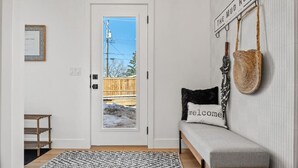 A clean, modern entryway with a white door, a bench with a 'welcome' pillow, a woven rug, and a wall-mounted sign reading 'THE MUD ROOM'. - The Duke | Spacious | Upscale | Close to DT (Calgary)
