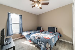 A bedroom with two beds, a ceiling fan, a window with curtains, and a desk with a computer.