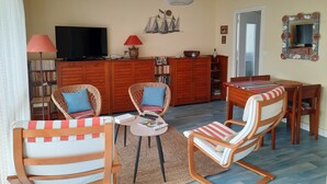 A living room with wicker chairs, a dining table, a TV, and a bookshelf. - 42m² overlooking the pine forest and near the sea (Les Sables-d'Olonne)