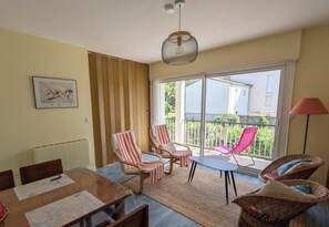 A room with a dining table, two striped armchairs, a wicker chair, a small table, and a balcony with a view.