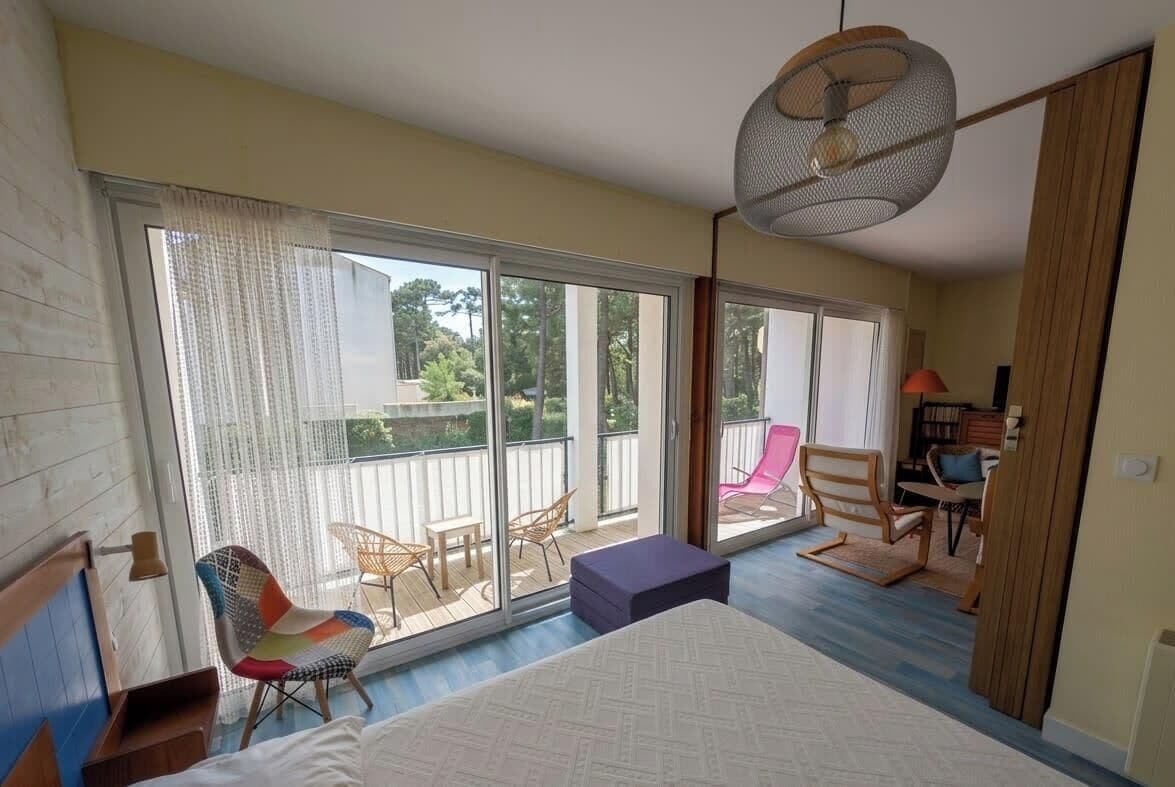 A bedroom with a large bed, a ceiling fan, a balcony with chairs, and a view of the outdoors.