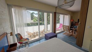 A bedroom with a large bed, a ceiling fan, a balcony with chairs, and a view of the outdoors. - 42m² overlooking the pine forest and near the sea (Les Sables-d'Olonne)