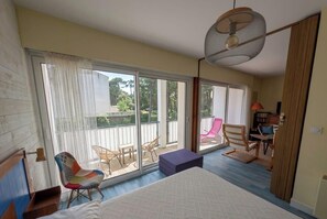 A bedroom with a large bed, a ceiling fan, a balcony with chairs, and a view of the outdoors.