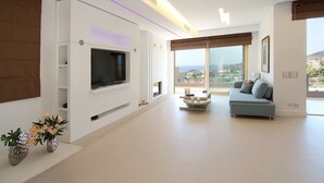 A modern living room with a flat-screen TV mounted on a white wall, a light blue sofa, a coffee table, and a view of the city through large windows. - Villa Nelmar, Luxury Villas in Anavyssos (Athens)