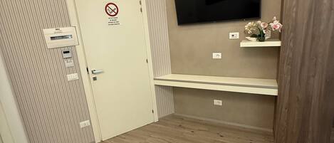 A bedroom with a bed, a TV mounted on the wall, a door with a no smoking sign, and a small shelf with flowers.