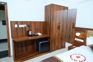 Quarto de hotel com guarda-roupa de madeira, pequena cozinha, mesa de cabeceira com abajur e cama com travesseiro decorativo.