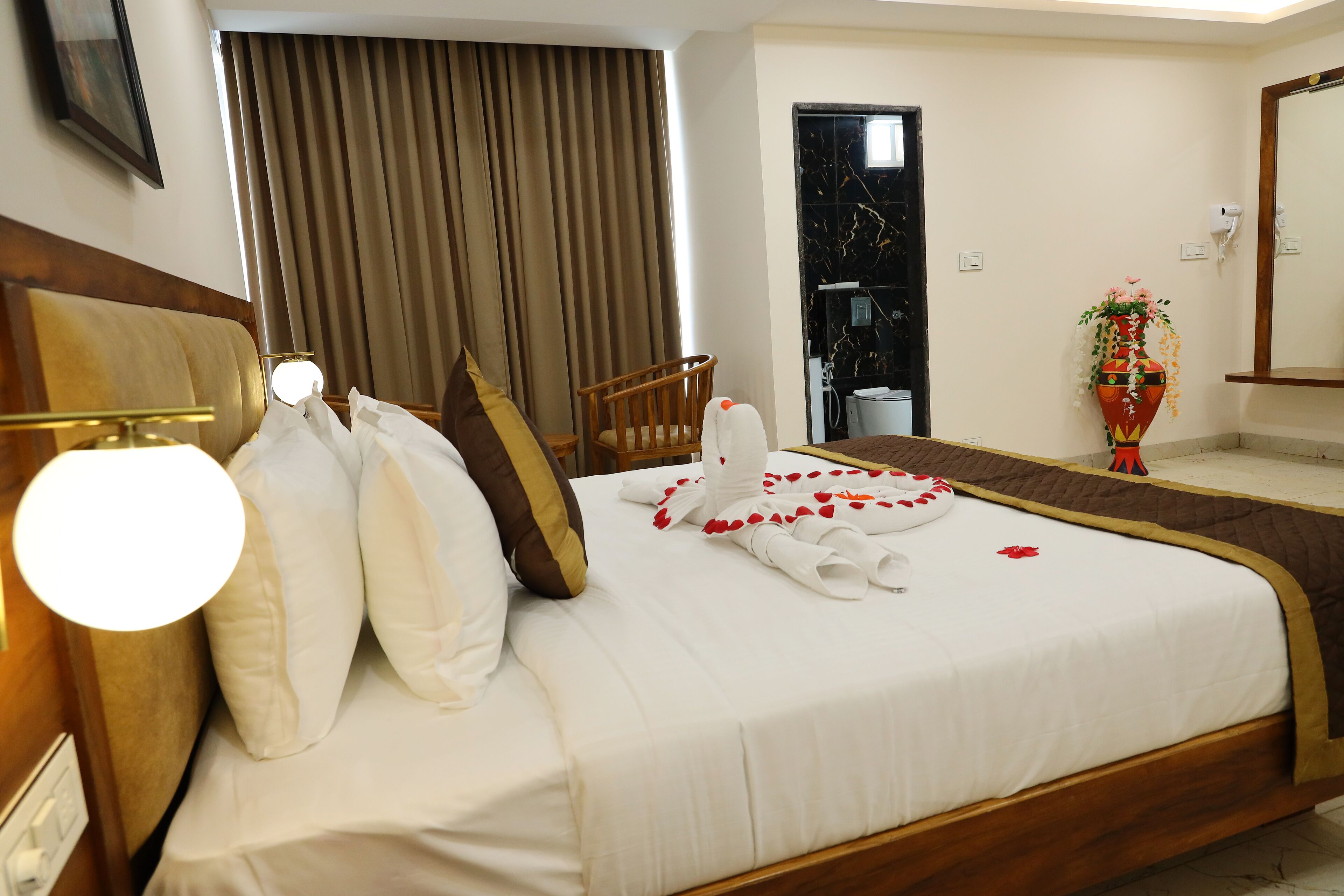 A hotel room with a neatly made bed, a wooden headboard, a wooden chair, a potted plant, and a bathroom visible through an open door.