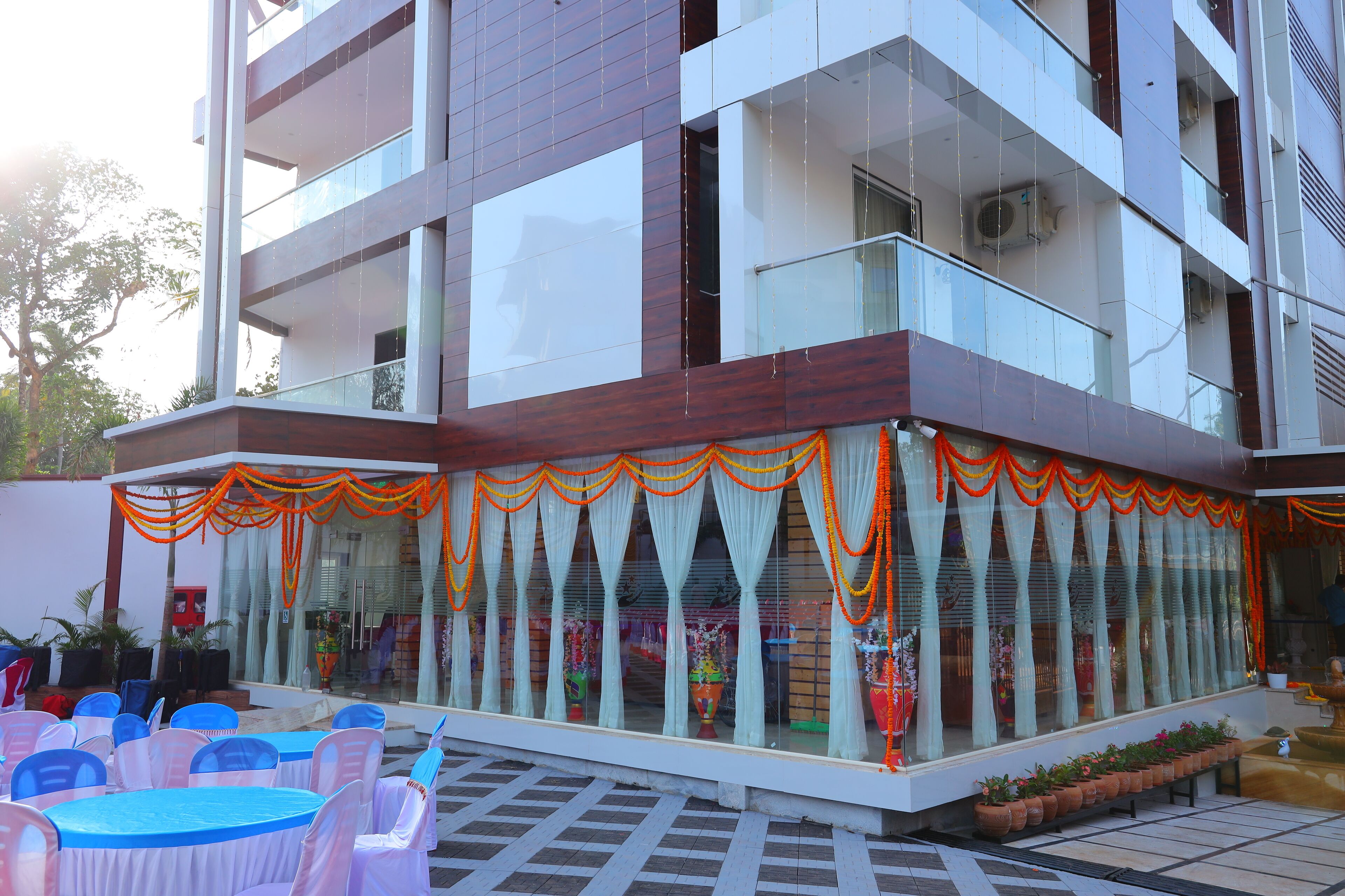 A modern building with a glass facade, outdoor seating area with blue and white tables, and potted plants along the walkway.