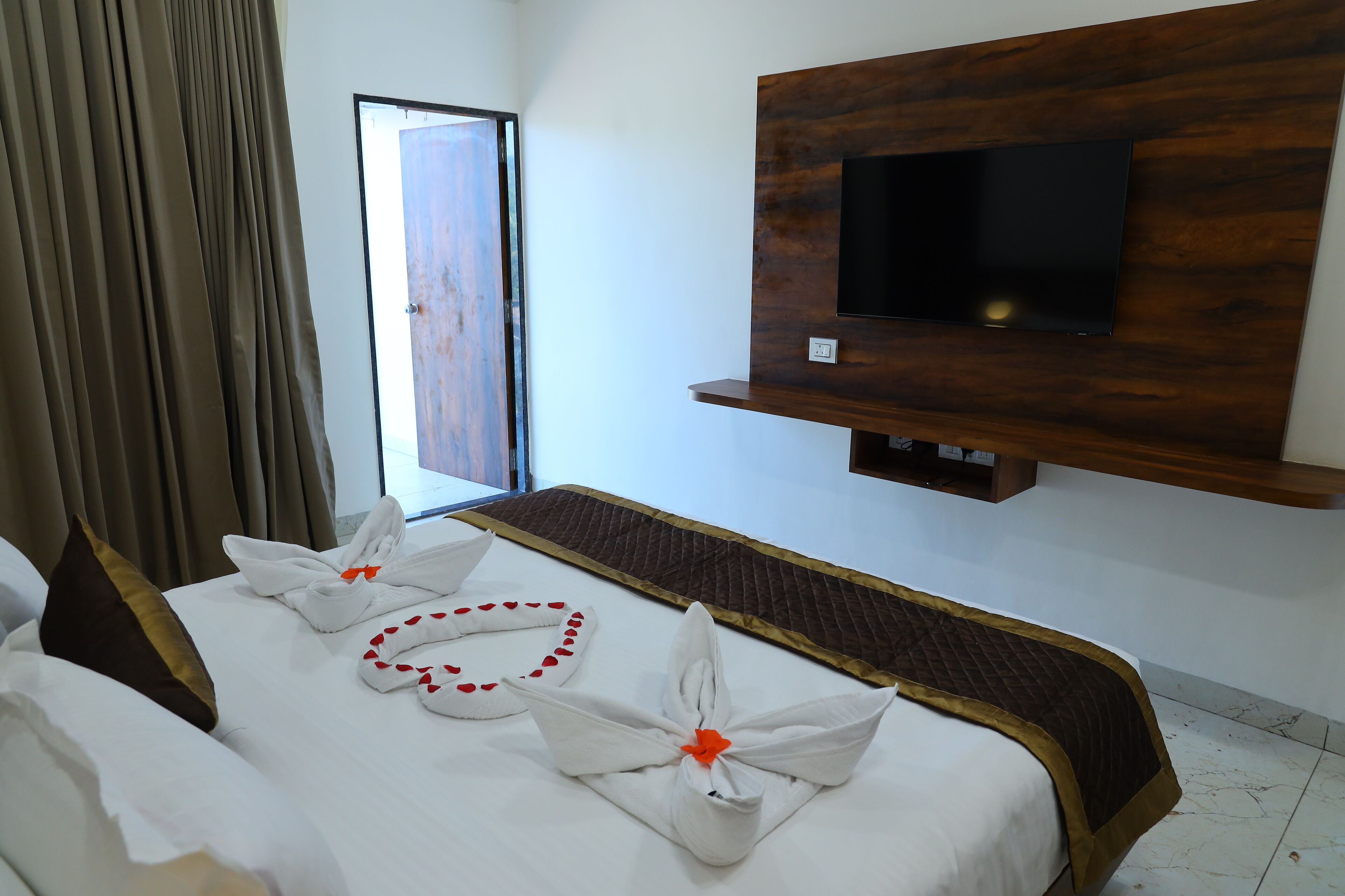 A hotel room with a bed, a TV mounted on a wooden panel, and a window with curtains.