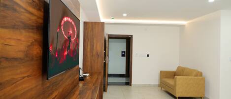 A reception area with a wooden reception desk, a modern wall-mounted TV, a beige sofa, and a glass door leading to a hallway.