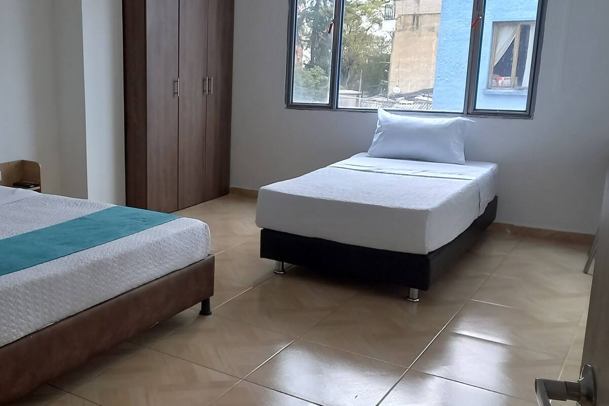 A hotel room with two beds, a wooden headboard, a teal bedspread, and a window with a view of buildings.