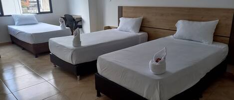 A hotel room with three beds, each with white bedding and pillows. There is a wooden headboard on the right bed. The room has a tiled floor and a window on the left side.