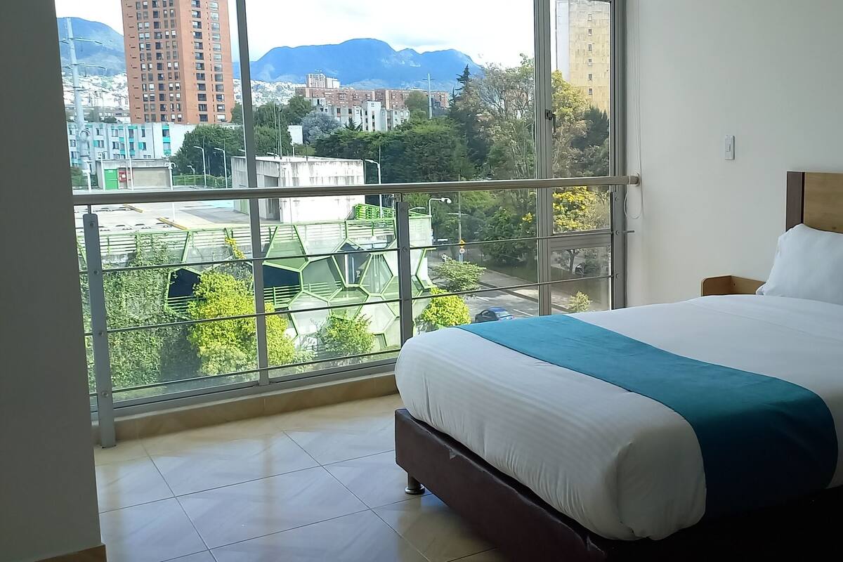 A hotel room with a large bed, a wooden headboard, and a view of a cityscape through a large window.