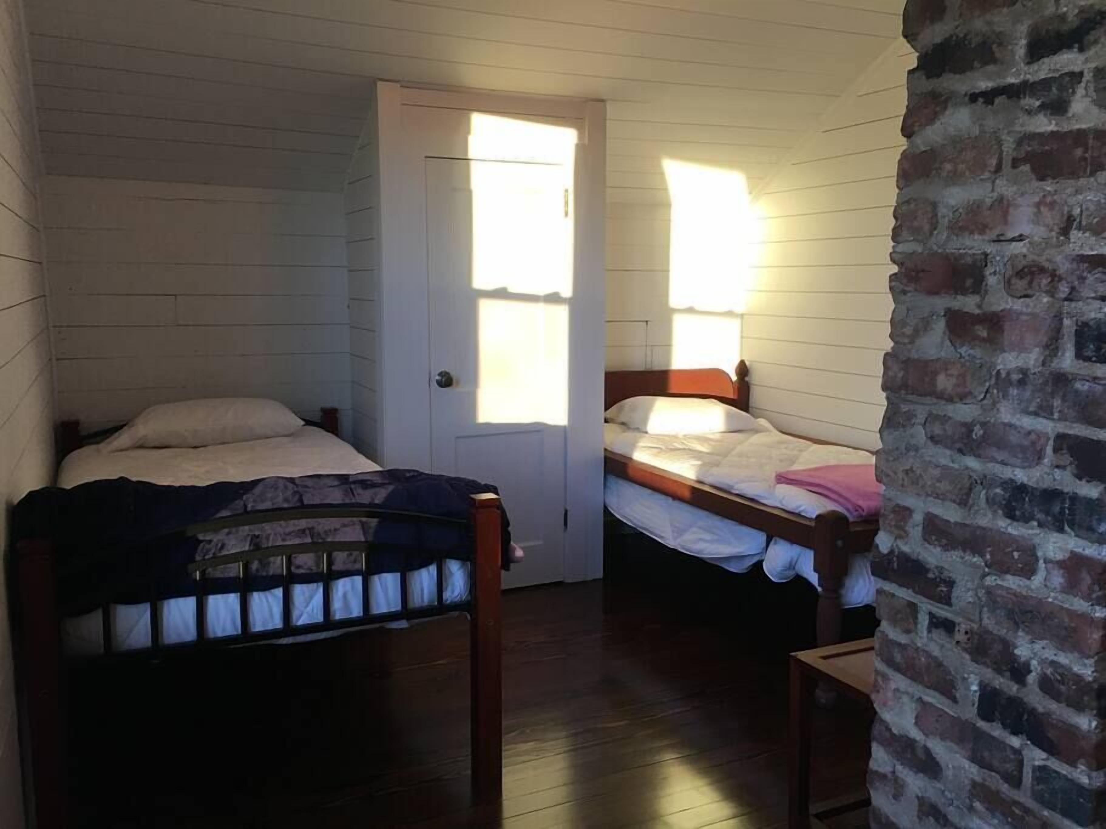 Une petite pièce chaleureuse avec deux lits en bois, un mur en briques et une porte blanche.