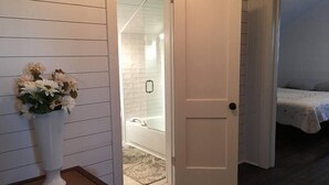 Habitació amb una porta blanca, una taula petita amb un ram de flors, una zona de banyera/dutxa i un llit amb un edredó estampat.