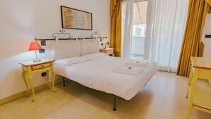 Habitación de hotel con cama, mesita de noche, lámpara, ventana con cortinas y una obra de arte colgada en la pared.