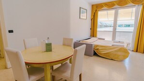 Une chambre d’hôtel avec une table ronde, deux chaises, un lit recouvert d’une couverture jaune, une fenêtre avec des rideaux jaunes et un tableau accroché au mur.