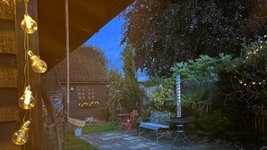 A stone patio with a metal bench and a table, surrounded by greenery and a house with a visible window. - Two Storey Annex (Cambridge)