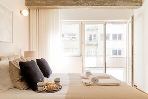 Una camera d'albergo con un letto, cuscini, un comodino con una lampada, una finestra vista edifici e asciugamani a terra.