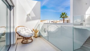 A balcony with a wicker chair, a glass railing, and a view of buildings and palm trees.