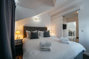 Hotelkamer met een groot bed, twee nachtkastjes met lampen, een modern hoofdeinde en uitzicht op de eetruimte door een geopende deur.