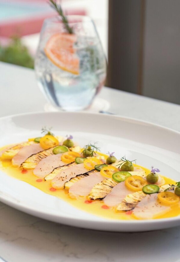 Un plato blanco con pescado en rodajas, salsa amarilla y adornos verdes sobre una superficie de mármol.